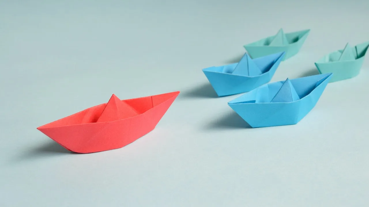 A red origami paper boat leading a group of blue paper boats